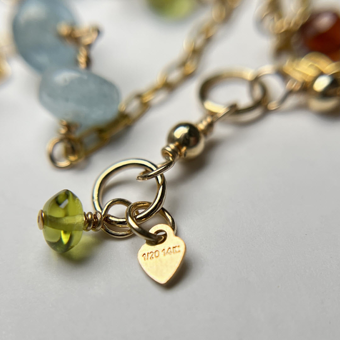 Close-up of a multicolor multi gemstone asymmetrical station necklace in 14K gold-fill staple chain showing quality tag and peridot dangle